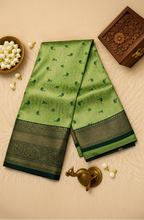 A folded green silk saree with a wide black and gold border and small green floral motifs, displayed on a beige faux fur surface with decorative props including a peacock figurine, white flowers, and a wooden box.