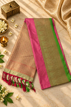 A folded Indian silk saree displayed on a marble surface with decorative props. The saree has a beige body with fine red and gold woven patterns, a vibrant pink border, and a green and gold zari-painted pallu.