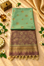 A folded silk saree displayed on a beige marble surface, featuring a light green upper body with gold sunburst motifs and a deep purple border with gold zari floral patterns and tassels. The saree is styled with white flowers, green leaves, a brass box, and a brass lamp.