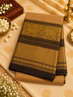 A folded beige and gold saree or fabric piece with ornate woven borders, displayed on a matching patterned cloth with decorative props including white flowers, a brass lamp, and a wooden box.