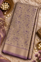 A folded beige and purple brocade saree with floral motifs and a metallic border, styled on a white faux fur background with decorative props.