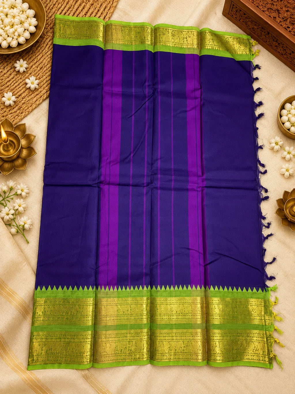 A purple and green silk saree with gold zari borders, laid flat on a beige fabric background decorated with brass lamps, white flowers, and a woven mat.