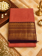 A folded terracotta-colored silk saree with an ornate gold and brown zari border, displayed on a beige patterned fabric alongside decorative brass lamps, a bowl of white flowers, and a carved wooden tray.